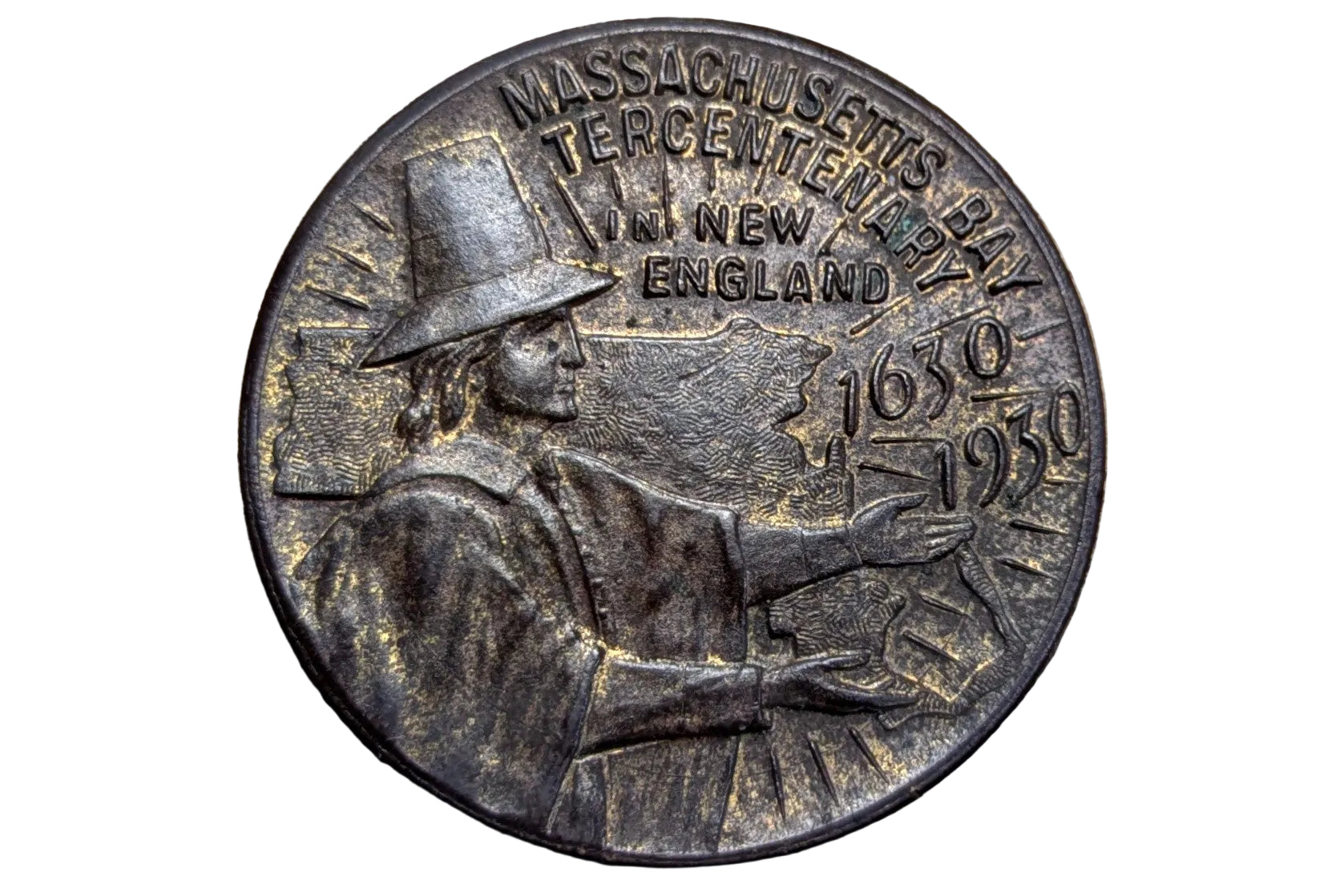 A round bronze commemorative coin of the Massachusetts Centennial 250th Anniversary of New England, featuring a profile of a Puritan figure and the years 1639 and 1939, displayed on a wooden surface.