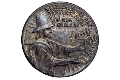 A round bronze commemorative coin of the Massachusetts Centennial 250th Anniversary of New England, featuring a profile of a Puritan figure and the years 1639 and 1939, displayed on a wooden surface.