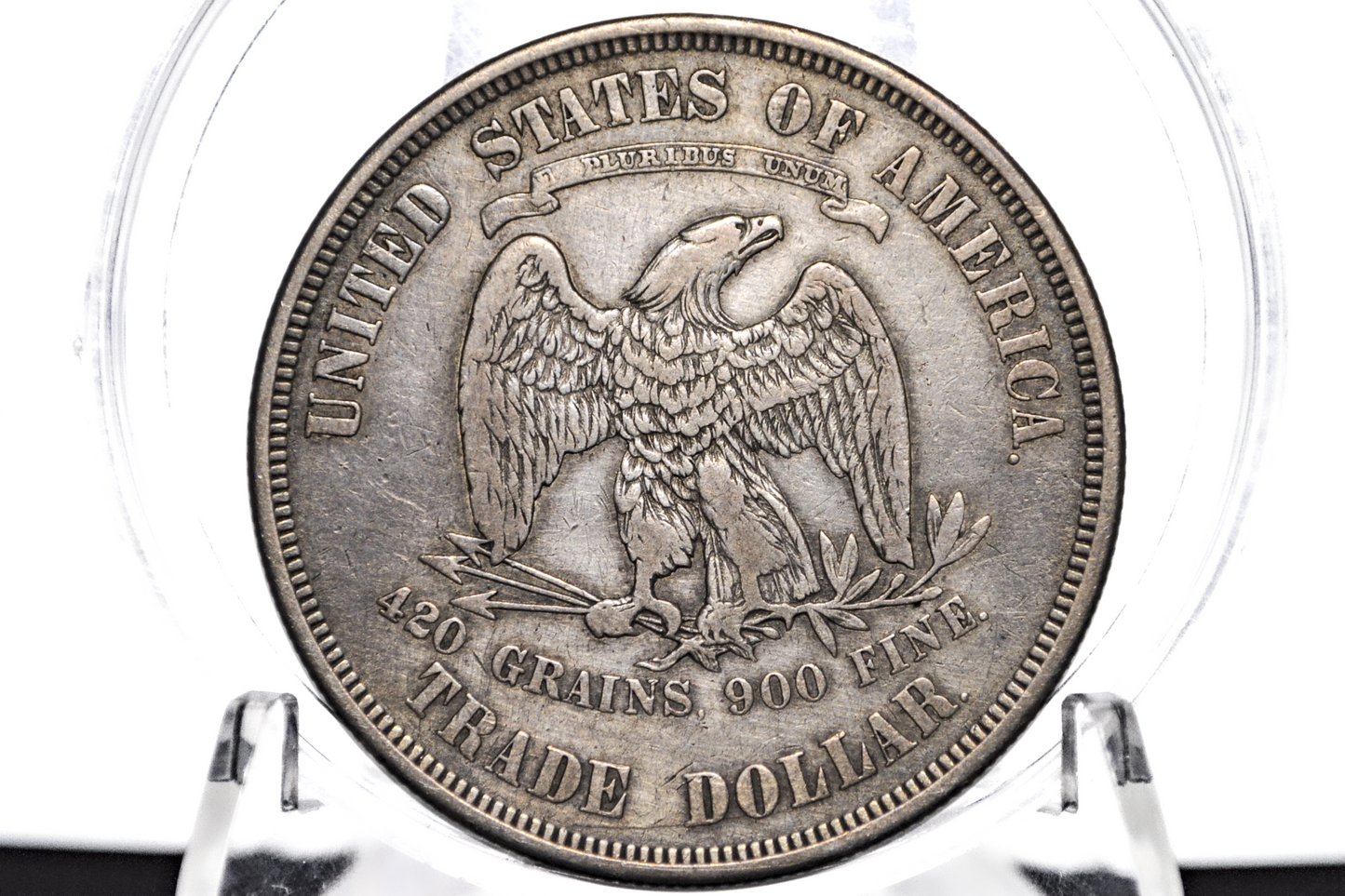 A silver-colored United States Trade Dollar coin sealed inside a clear plastic protective capsule, displayed on a clear stand against a white background.