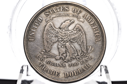 A silver-colored United States Trade Dollar coin sealed inside a clear plastic protective capsule, displayed on a clear stand against a white background.