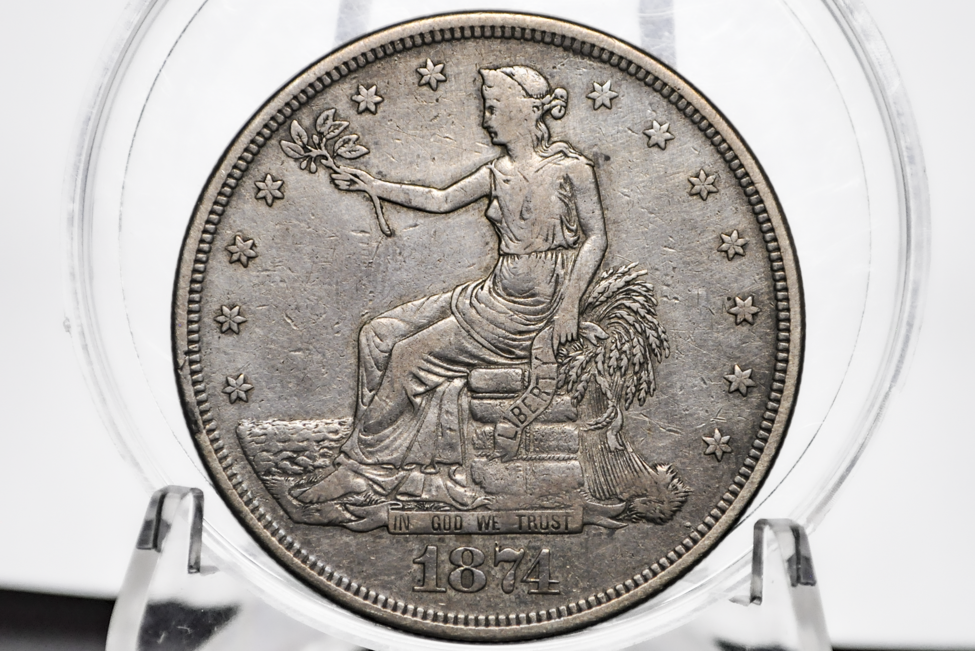 An 1874 United States Trade Dollar displayed inside a clear plastic grading holder, showing the seated female figure design on a silver-toned metal surface. Dated 1874
