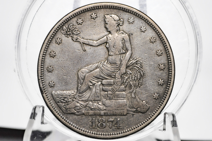 An 1874 United States Trade Dollar displayed inside a clear plastic grading holder, showing the seated female figure design on a silver-toned metal surface. Dated 1874