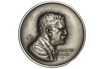A silver-toned commemorative coin featuring a profile bust of Theodore Roosevelt, displayed inside a clear plastic protective capsule on a light gray background.